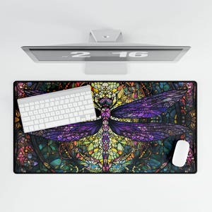 Può includere: Un tappetino per scrivania rettangolare con un design a libellula in stile vetrata colorata nei toni del viola, verde e giallo. Una tastiera e un mouse bianchi sono sul tappetino. Un monitor per computer è sopra il tappetino.