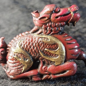 May include: A red and gold Chinese dragon figurine. The dragon is sitting on its haunches with its head turned to the side. The dragon is decorated with intricate details and has a gold finish.