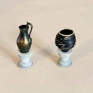 May include: Two miniature ceramic pots on white pedestals. The pot on the left is a green and gold pitcher. The pot on the right is black with white markings.