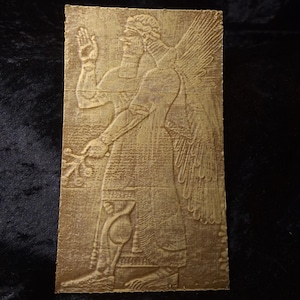 May include: A gold-coloured rectangular relief carving of a winged figure in ancient dress. The figure is raising one hand and holding a sprig. The carving has detailed textures and is set against a dark background.