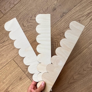 Può includere: Tre lunghe strisce rettangolari di legno chiaro non rifinito. Ogni striscia presenta un bordo decorativo smerlato su un lato, creando una serie di ritagli arrotondati a semicerchio. La venatura del legno è visibile sulle superfici piatte.