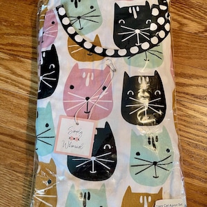May include: A white apron set with a repeating pattern of cat faces in black, pink, teal, and gold. The apron has a black and white polka dot strap and a tag that reads "Simply Whimsical". The packaging also includes the text "Crazy Cat Apron Set".