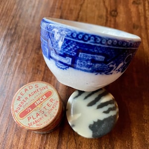 May include: A collection of vintage items on a wooden surface. Includes a small blue and white ceramic bowl, a round container of "MEADS RUBBER ADHESIVE 1/2 INCH PLASTER", and a black and white marbled stone.