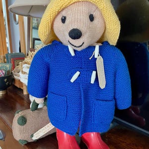 May include: A handmade Paddington Bear plush toy. The bear wears a blue coat, yellow hat, and red boots. It holds a small suitcase. The bear has a black nose and a tag attached to its coat.