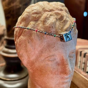 May include: A silver-toned headpiece with a small, square pendant featuring a figure in a pose. The headpiece has a delicate chain with colorful beads. The headpiece is displayed on a bust of a classical figure.