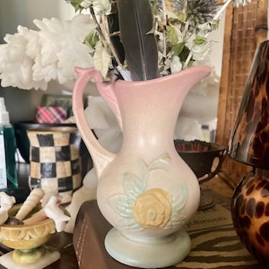 May include: A vintage pink ceramic pitcher with a handle, featuring a raised yellow rose design and light blue accents. The pitcher holds dried flowers and a dark feather. The pitcher is on a wooden surface.