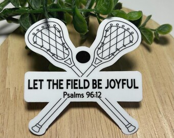 Może przedstawiać: Biała naklejka przedstawiająca dwa skrzyżowane kije do lacrosse z czarnym obrysem oraz małe czarne koło pośrodku. Pod kijami znajduje się prostokątny baner z czarnym napisem LET THE FIELD BE JOYFUL oraz odniesieniem Psalms 96:12.