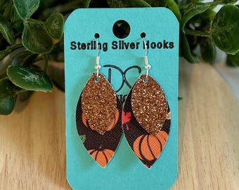 Może przedstawiać: Para kolczyków wiszących z nadrukiem dyni i złotym brokatem. Kolczyki znajdują się na turkusowej kartce z napisem "Sterling Silver Hooks".