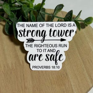 May include: A white sticker with black text. The text reads "THE NAME OF THE LORD IS A strong tower" with an arrow graphic. Below, it says "THE RIGHTEOUS RUN TO IT AND are safe" and "PROVERBS 18:10". The sticker is on a wooden surface.
