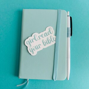 Pu&ograve; includere: Un taccuino azzurro con chiusura elastica verticale e una penna bianca attaccata sul lato. Un adesivo bianco sulla copertina riporta il testo "girl read your bible" in corsivo azzurro. Un piccolo segnalibro a nastro sporge dalla parte inferiore.