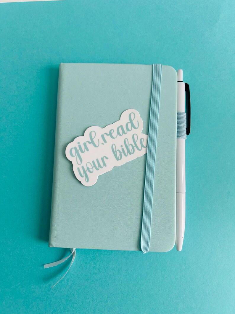 Pu&ograve; includere: Un taccuino azzurro con chiusura elastica verticale e una penna bianca attaccata sul lato. Un adesivo bianco sulla copertina riporta il testo "girl read your bible" in corsivo azzurro. Un piccolo segnalibro a nastro sporge dalla parte inferiore.
