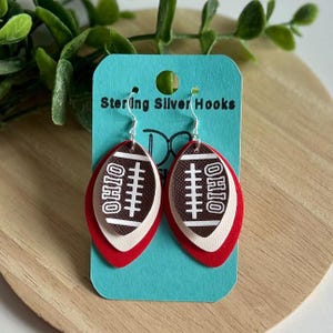 May include: Sterling Silver Hooks earrings. These earrings feature a football design with the word "OHIO" and white stitching details. The earrings are layered with red, white, and brown colors. The earrings are hanging on a blue card.