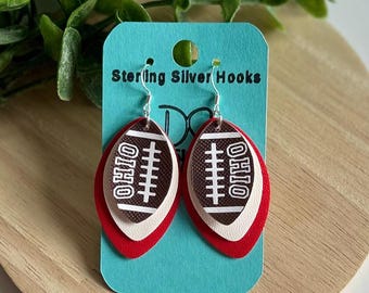Può includere: Orecchini Sterling Silver Hooks. Questi orecchini presentano un design a forma di pallone da football con la scritta "OHIO" e dettagli di cucitura bianchi. Gli orecchini sono stratificati con colori rosso, bianco e marrone. Sono appesi a una carta blu.