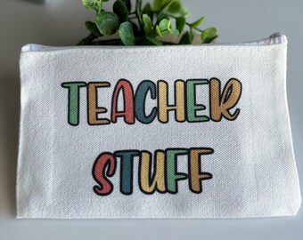 Może przedstawiać: Biała płócienna kosmetyczka zapinana na zamek z napisem "TEACHER STUFF" wydrukowanym kolorowymi literami. Kosmetyczka jest prostokątna i ma biały zamek błyskawiczny. W tle widoczna jest zieleń.