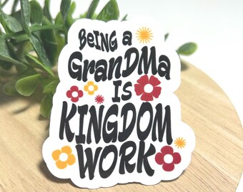 Może przedstawiać: Biała naklejka z wyciętym konturem, zawierająca czarny napis "Being a GranDMA is KINGDOM WORK" pogrubioną, falistą czcionką. Wzór ozdobiony jest małymi, kolorowymi ilustracjami kwiatów w odcieniach czerwieni, pomarańczu i żółci oraz ikoną słońca.