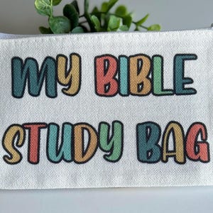 Pu&ograve; includere: Una pochette con cerniera in tela bianca con il testo MY BIBLE STUDY BAG stampato in un carattere multicolore di ispirazione retr&ograve;. Le lettere si alternano tra tonalit&agrave; tenui di blu, giallo, rosso e verde, ciascuna con un bordo nero.