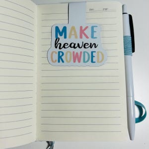 Pu&ograve; includere: Un segnalibro magnetico con il testo MAKE heaven CROWDED in lettere blu, rosa e gialle, fissato sulla parte superiore di un taccuino aperto a righe. Una penna bianca con impugnatura testurizzata azzurra &egrave; posizionata verticalmente a destra.