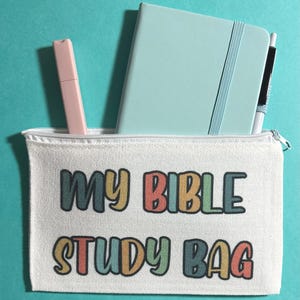 Pu&ograve; includere: Una pochette con cerniera in tela bianca con la scritta "MY BIBLE STUDY BAG" stampata in lettere colorate e arrotondate. All'interno della pochette si trovano un taccuino azzurro, un evidenziatore rosa pallido e una penna bianca e nera su sfondo verde acqua.