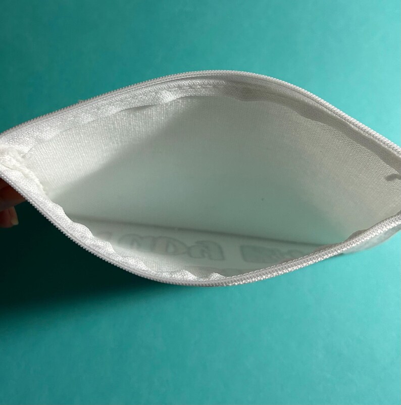 Pu&ograve; includere: Una pochette in tessuto bianco con cerniera e apertura semicircolare. L'interno &egrave; foderato con materiale bianco e l'esterno presenta una cerniera bianca lungo il bordo superiore. Visualizzazione da una prospettiva dall'alto.