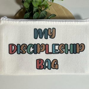 Pu&ograve; includere: Una pochette rettangolare con cerniera in tela color bianco sporco con il testo MY DISCIPLESHIP BAG stampato in un carattere retro multicolore. Le lettere alternano tonalit&agrave; tenui di rosso, blu, verde e marrone con un contorno scuro.