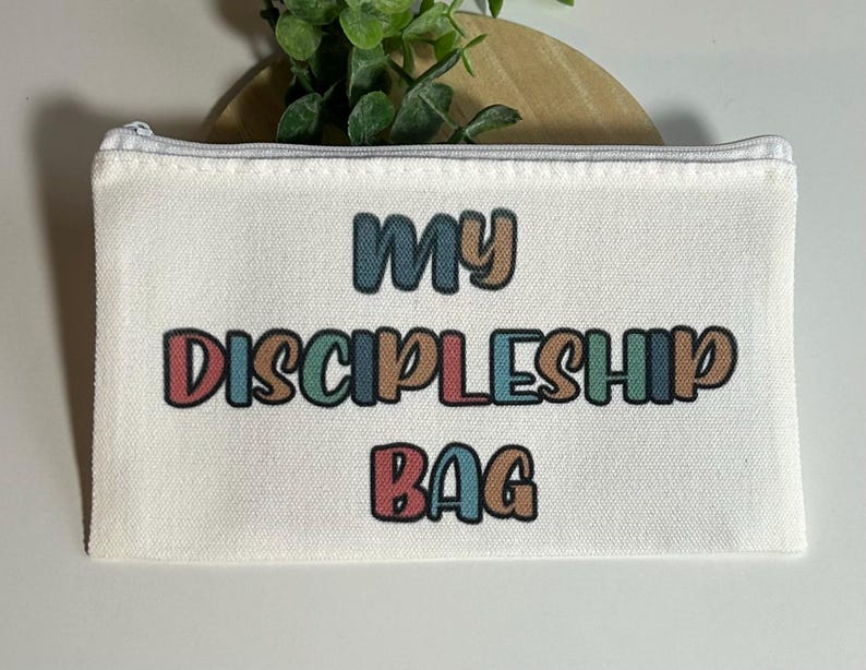 Pu&ograve; includere: Una pochette rettangolare con cerniera in tela color bianco sporco con il testo MY DISCIPLESHIP BAG stampato in un carattere retro multicolore. Le lettere alternano tonalit&agrave; tenui di rosso, blu, verde e marrone con un contorno scuro.