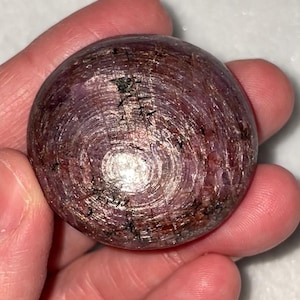May include: A polished, round gemstone with a deep reddish-purple hue. The stone has a swirling, textured pattern with hints of gold and black inclusions. The gemstone is held in a hand, showcasing its size and shape.