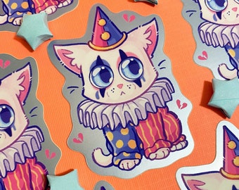 May include: A silver holographic sticker featuring a cartoon cat dressed as a clown. The cat has a white face with black eyeliner, blue eyes, and a pink nose. It is wearing a purple and yellow jester hat, a purple ruffle collar, and a pink and yellow striped costume with blue polka dots. The sticker has a distressed, broken heart design in the background.