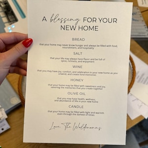 May include: A white card with black text that reads "A blessing for your new home." The card lists items like bread, salt, wine, honey, olive oil, and candle, with a blessing for each item. The card is signed "Love, The Waldmanns."