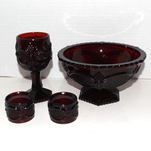 May include: A set of ruby red glass tableware, including a goblet, a pedestal bowl, and two small round containers. The pieces have a textured, patterned design. The bowl is approximately 8 inches in diameter.