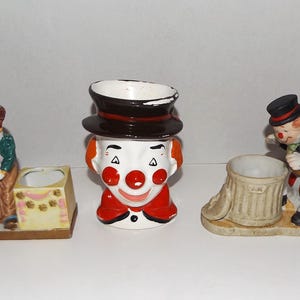 May include: Three ceramic clown figurines. One is a clown head with a top hat, another is a clown figure with a small box, and the third is a clown figure with a trash can. The clowns have red noses and painted details.