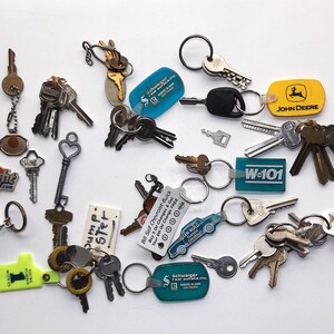 May include: A collection of keys and keychains of various shapes and sizes. Some keychains have logos, including "BlueLight", "John Deere", and "W-101". The keys are made of metal and are arranged on a white surface.