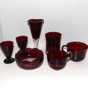 May include: A collection of six deep red glass items including two stemmed goblets, a tall fluted glass with a clear base, a wide scalloped bowl, a cylindrical glass, and a handled mug. The bowl and mug feature faint embossed text on their sides.