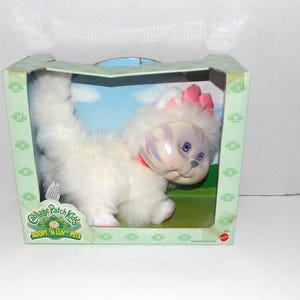 May include: A Cabbage Patch Kids Adopt 'N Luv Pets kitten doll in its original box. The white kitten has a pink bow, pink accents, and blue eyes. The box is green with the Cabbage Patch Kids logo.