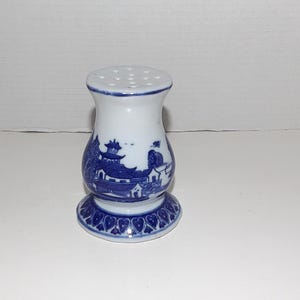 May include: A white ceramic shaker with a blue willow-style pattern featuring pagodas and trees. The shaker has a flared base decorated with a repeating heart-shaped motif and a top surface perforated with multiple small holes for dispensing.