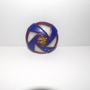 May include: A vintage Simon game console. The game has a circular design with a blue and purple exterior. The center features the word "Simon" in gold lettering. The game has four colored buttons and a speaker.