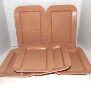 May include: A set of five rectangular trays in a solid muted peach color. Each tray features a smooth, glossy finish with rounded corners and a raised border edge. The trays are arranged in a stacked formation with two standing upright.
