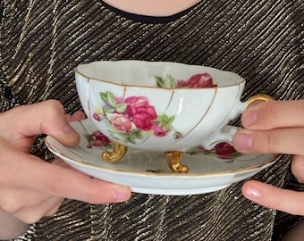 Puede incluir: Una taza de té y un platillo blancos con un diseño floral. La taza tiene tres patas doradas y un borde dorado. El platillo tiene un borde dorado y un diseño floral.