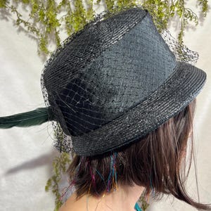 May include: A black hat with a brim and a decorative green feather. The hat features a fine black netting and a textured surface. The hat is a stylish accessory.