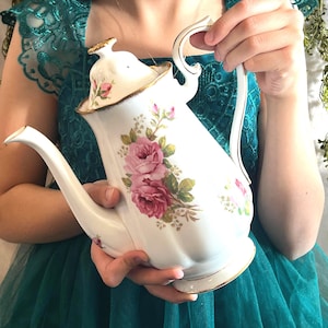 Puede incluir: Tetera de porcelana blanca con un diseño floral de rosas en rosa y verde. La tetera tiene un ribete dorado y un pico y asa curvos. La tapa también está decorada con rosas. Un artículo clásico para la hora del té.