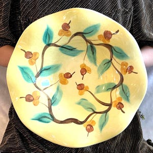 May include: A yellow ceramic serving dish with a hand-painted design. The design features a brown branch with green leaves and yellow and brown berries. The dish has an irregular, wavy edge.