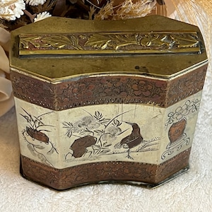 May include: An antique, multi-sided metal box with a hinged lid. The box features a decorative floral and bird design in brown, white, and gold tones. The top and bottom edges have ornate embossed patterns. The box is likely used for storage.