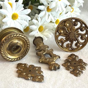 May include: A collection of brass hardware components including a circular knob with a rope-textured edge, a vertical drop pull with an ornate design, a circular decorative medallion with openwork scroll patterns, and two small diamond-shaped plates.