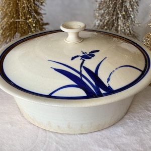 May include: A white ceramic serving dish with a blue and brown rim and a blue floral design on the lid. The dish has a knob on the lid.