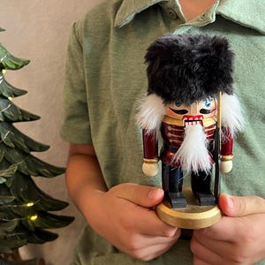 May include: A wooden nutcracker figure wearing a tall black faux fur hat, a red jacket with gold trim, and dark blue trousers. The figure has a white faux fur beard, painted facial features, and stands on a round gold base, holding a thin black staff.