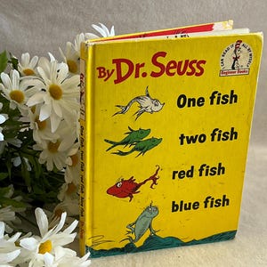 Pode incluir: Um livro amarelo de Dr. Seuss intitulado "One Fish, Two Fish, Red Fish, Blue Fish" com ilustrações de peixes. Margaridas brancas com centros amarelos circundam o livro.