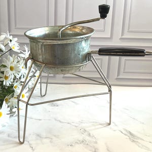May include: A vintage metal food mill featuring a silver-toned basin with a rotating crank handle, a black plastic grip, and a sturdy wire stand. The kitchen tool is designed for pureeing or straining food items and sits on a thin metal frame.
