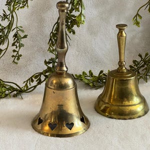 May include: Two antique brass bells. The larger bell has heart-shaped cutouts near the base. Both bells have a tarnished, aged appearance. Greenery is draped in the background, adding a touch of nature to the scene.