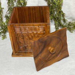 May include: A wooden flour container with a lid. The container is rectangular with carved details, including the word "FLOUR" and a tropical scene. The lid is square with a circular design and a wooden knob.
