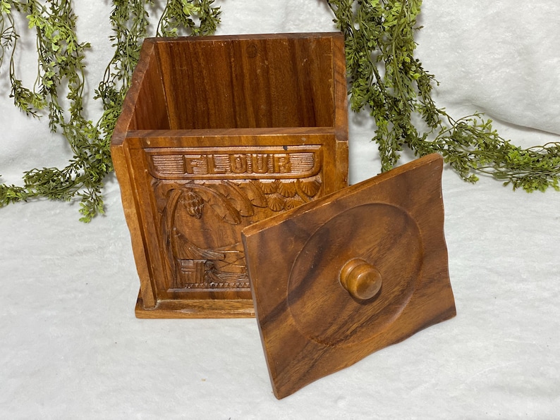May include: A wooden flour container with a lid. The container is rectangular with carved details, including the word "FLOUR" and a tropical scene. The lid is square with a circular design and a wooden knob.