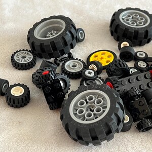 Op de afbeelding: Een verzameling zwarte speelgoedbanden in verschillende maten, sommige met grijze of gele velgen. Er zijn ook verschillende zwarte Lego-stenen aanwezig, sommige met rode accenten. De banden liggen verspreid op een wit oppervlak.